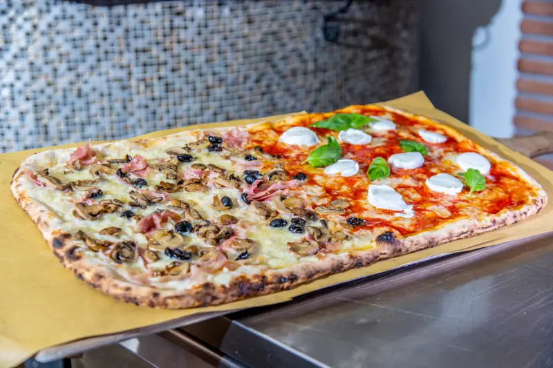 Pizza al mezzo metro a due gusti su carta da forno: metà bianca con funghi, prosciutto cotto e olive nere, metà rossa con mozzarella e basilico
