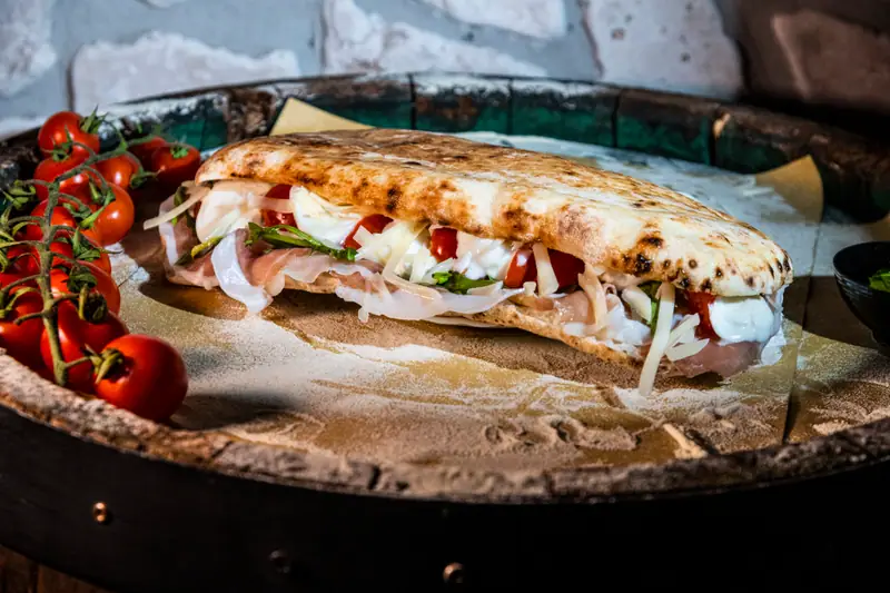 Panuozzo farcito con prosciutto crudo, mozzarella, pomodorini, rucola e scaglie di formaggio, su una botte infarinata accanto a pomodorini a grappolo