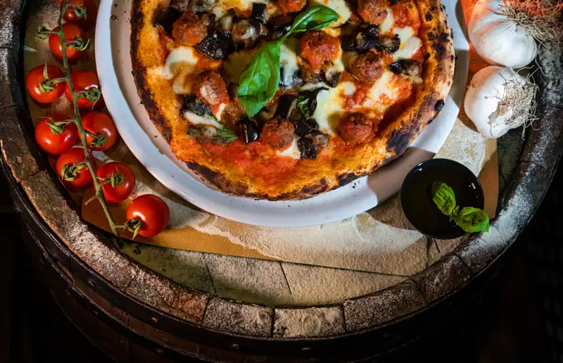 Pizza rossa con mozzarella, melanzane, pezzetti di carne e basilico su un piatto bianco, sopra una botte infarinata con pomodorini e aglio